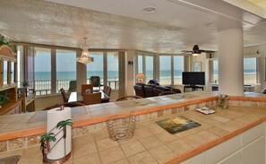 A spacious living area with a large window overlooking the beach, a ceiling fan, and a television.