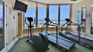Une salle de remise en forme équipée de tapis de course, d’un vélo d’appartement et d’un téléviseur. De grandes fenêtres offrent une vue sur la plage et l’océan.