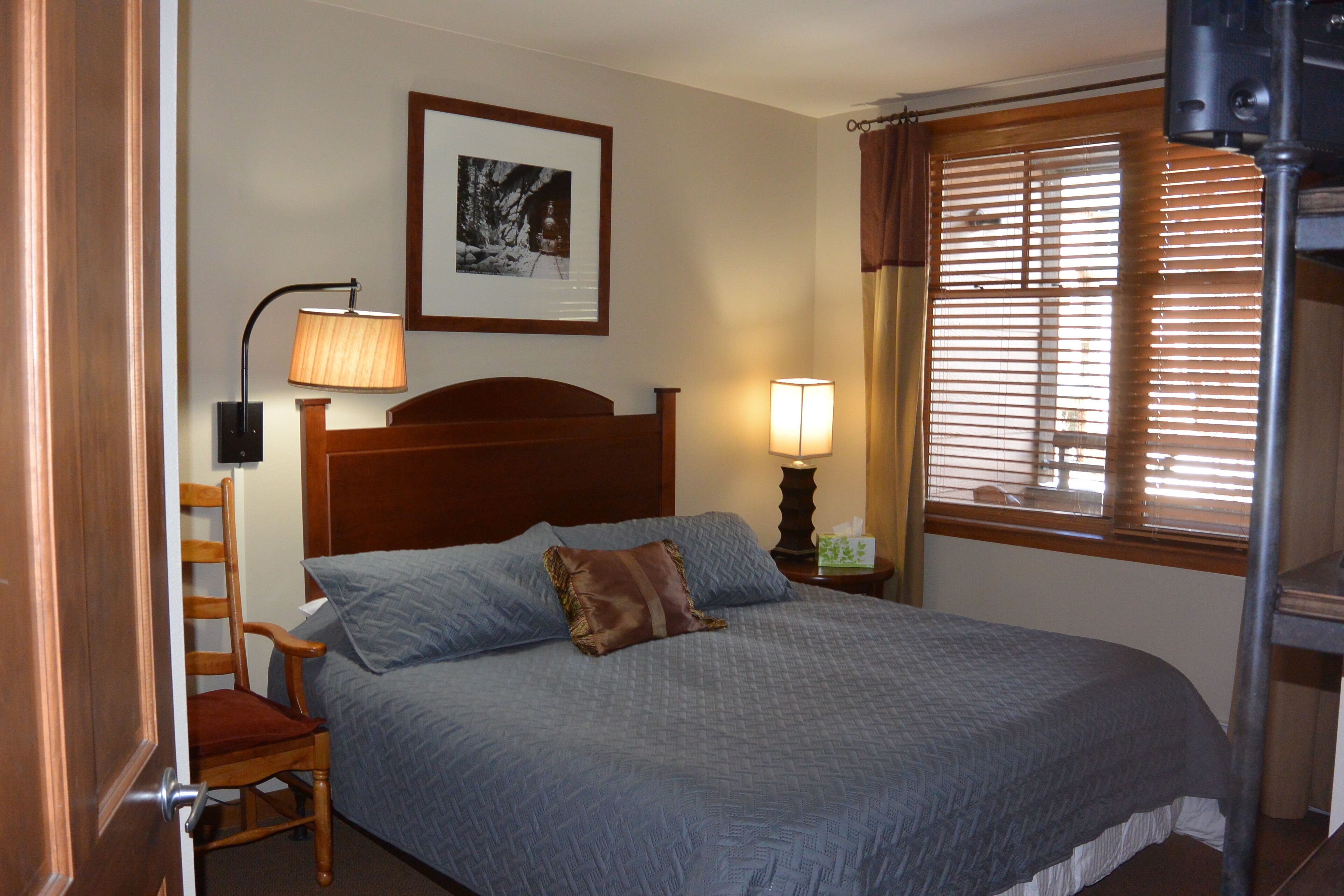 Cama bem arrumada com cabeceira, uma cadeira de madeira, mesa de cabeceira com abajur, quadro na parede e janela com persianas.