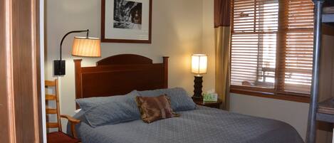 Una cama bien hecha con cabecera, una silla de madera, una mesita de noche con lámpara, un cuadro en la pared y una ventana con persianas.