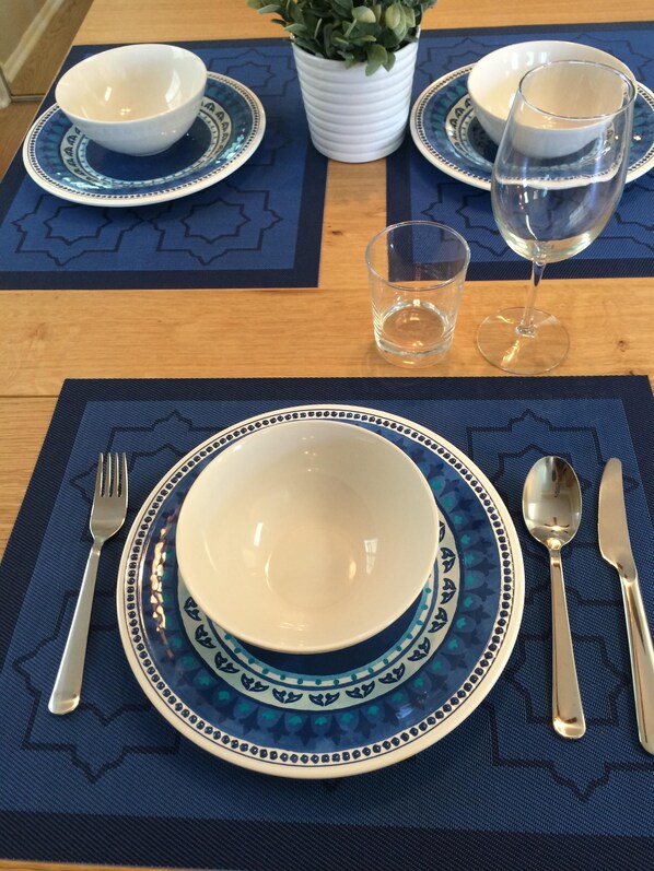 A dining table set with white plates, blue patterned table mats, a white bowl, a glass of water, and cutlery.