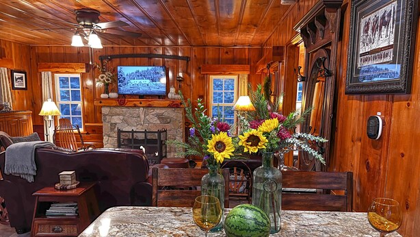 A cozy cabin interior with a stone fireplace, wooden walls, a television, and a dining table set with flowers and fruit. - Historic Hershey Cabin, Most 10 Star Reviews, Romantic, Just for 2, Immaculate (Lake Arrowhead)