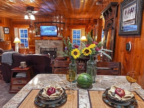 A cozy cabin interior with a stone fireplace, wooden walls, a television, and a dining table set with flowers and fruit. - Historic Hershey Cabin, Most 5 Star Reviews, Romantic, Just for 2, Immaculate (Lake Arrowhead)
