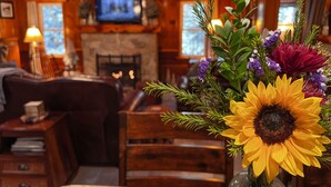 A cozy living room with a fireplace, a television mounted above it, and a floral arrangement on a table. - Historic Hershey Cabin, Most 10 Star Reviews, Romantic, Just for 2, Immaculate (Lake Arrowhead)