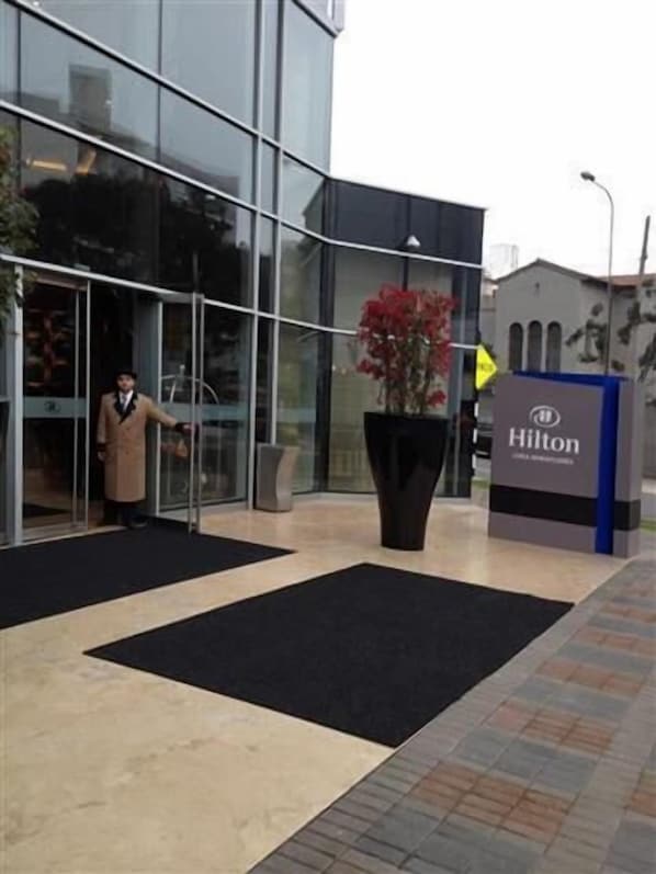 A modern hotel entrance with a glass facade, a black mat leading to the door, and a large potted plant with red flowers.