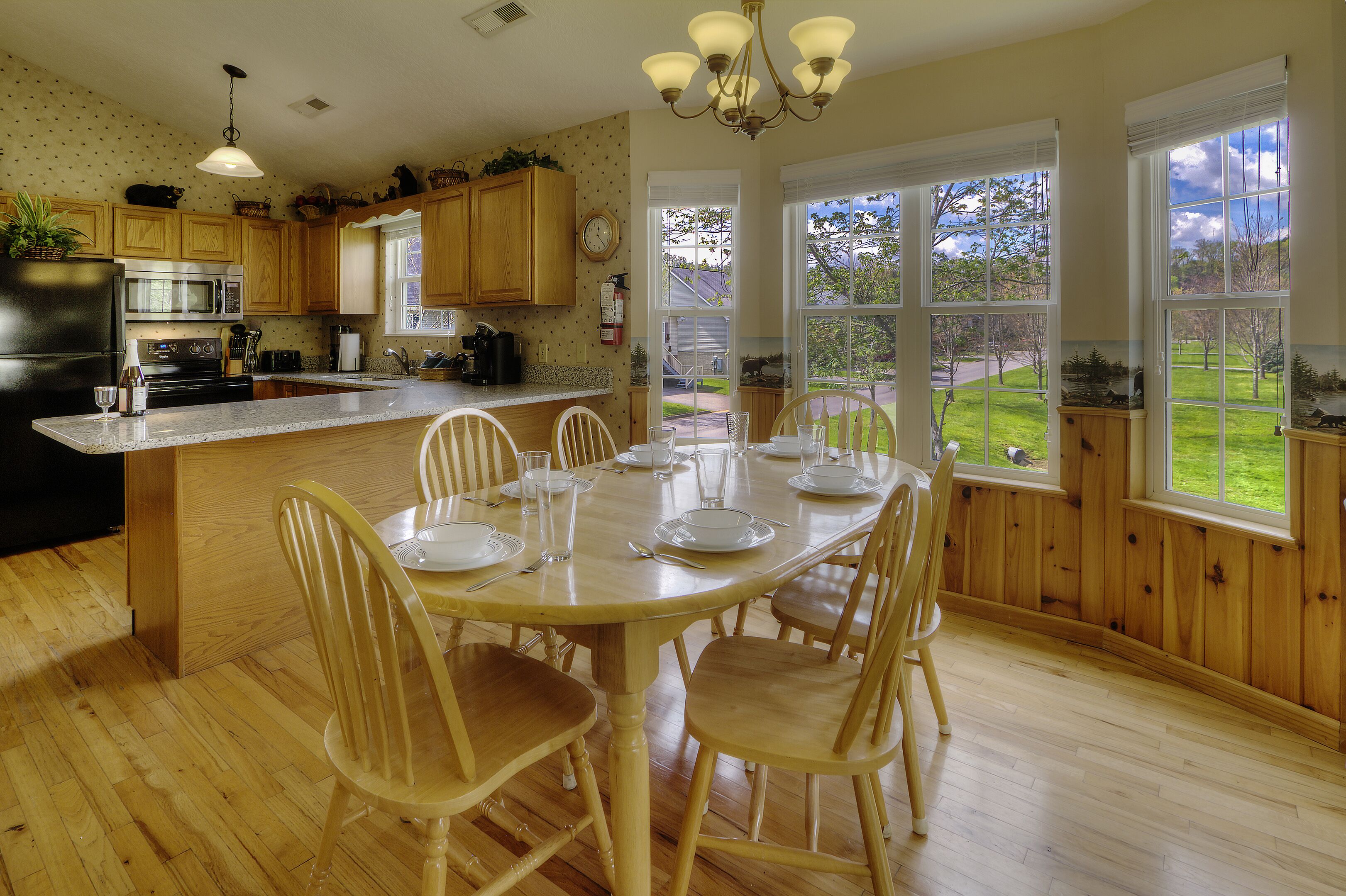 A kitchen with wooden cabinets, a dining table set for four, and large windows offering a view of the outdoors.