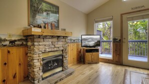 A stone fireplace with a television in front, wooden cabinets, and a large window with a view of trees. - Amy's Waterfront Blessing w/ Hot Tub, Arcade , & Fishing 2 King Master Suites (Pigeon Forge)