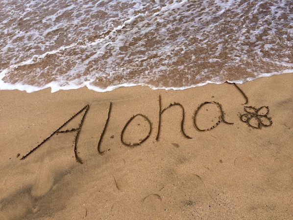 Uma praia com a palavra 'Aloha' e um desenho de flor na areia.