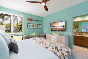 A bedroom with a bed, a ceiling fan, a window with blinds, a painting, and a bathroom with a mirror and sink. - 5 Star Ocean View, Beach House Chic, Click the Video tour below! (Wailea)