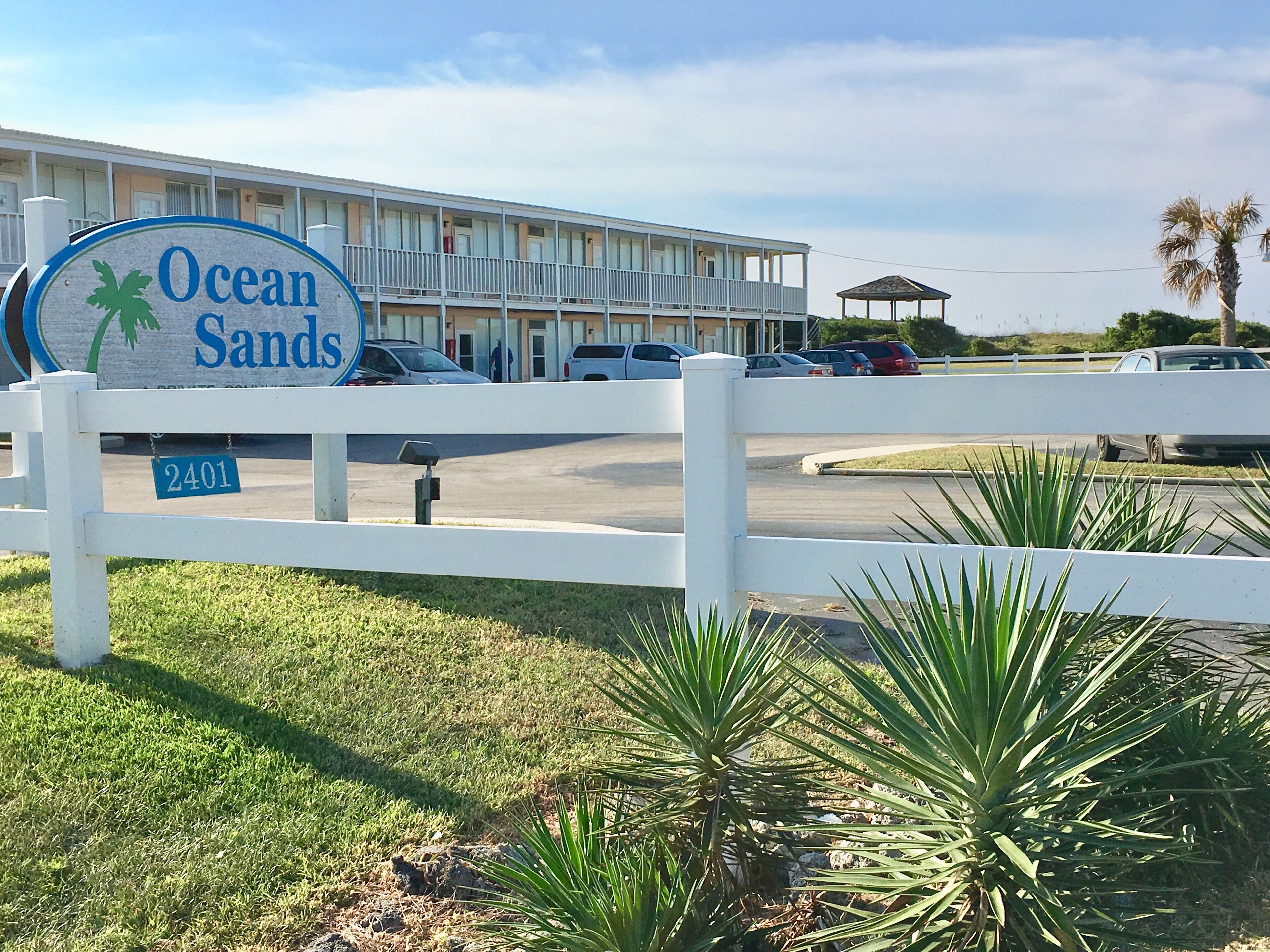 Een motel genaamd Ocean Sands, met een witte hek, een bord met palmbomen en een parkeerplaats.