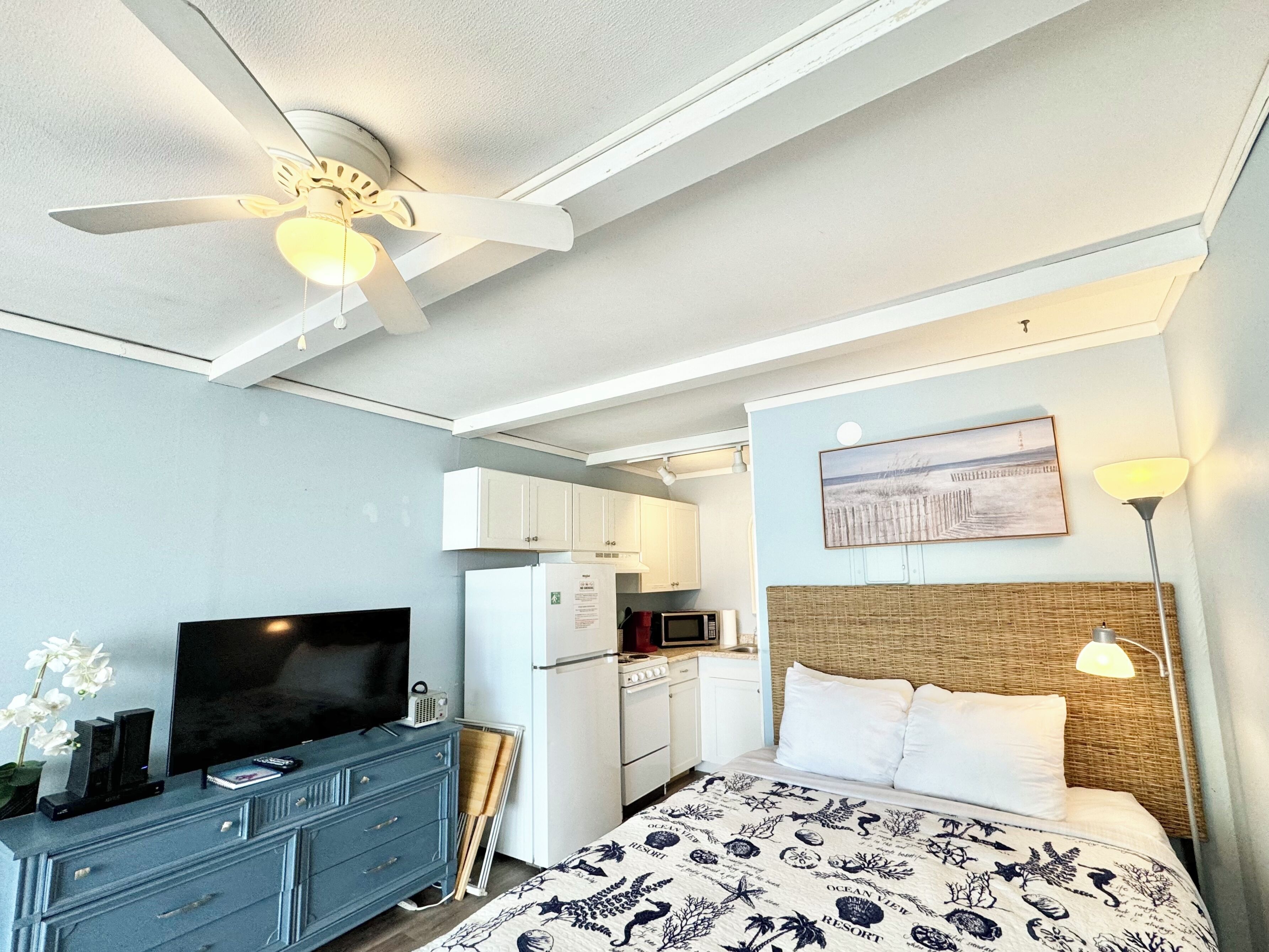 A hotel room with a bed, a TV, a ceiling fan, a microwave, and a kitchenette.