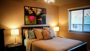 A neatly made bed with a quilted cover, two bedside lamps, and a wall-mounted artwork above it. - The 8th Street Retreat Looks Booked it may not be Call Us (Carbondale)