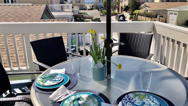 Ein runder Glastisch gedeckt mit Tellern, Besteck und einer Blumenvase auf einem Balkon mit Blick auf eine Wohngegend.