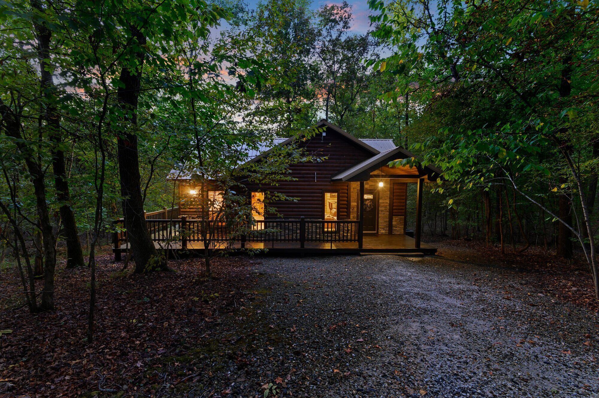 A rustic cabin with a wooden exterior, a porch, and a gravel driveway surrounded by trees.