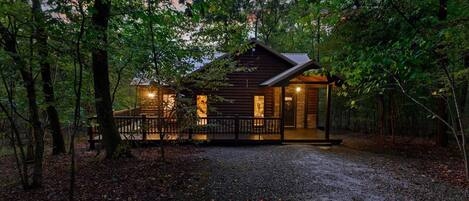 A rustic cabin with a wooden exterior, a porch, and a gravel driveway surrounded by trees.