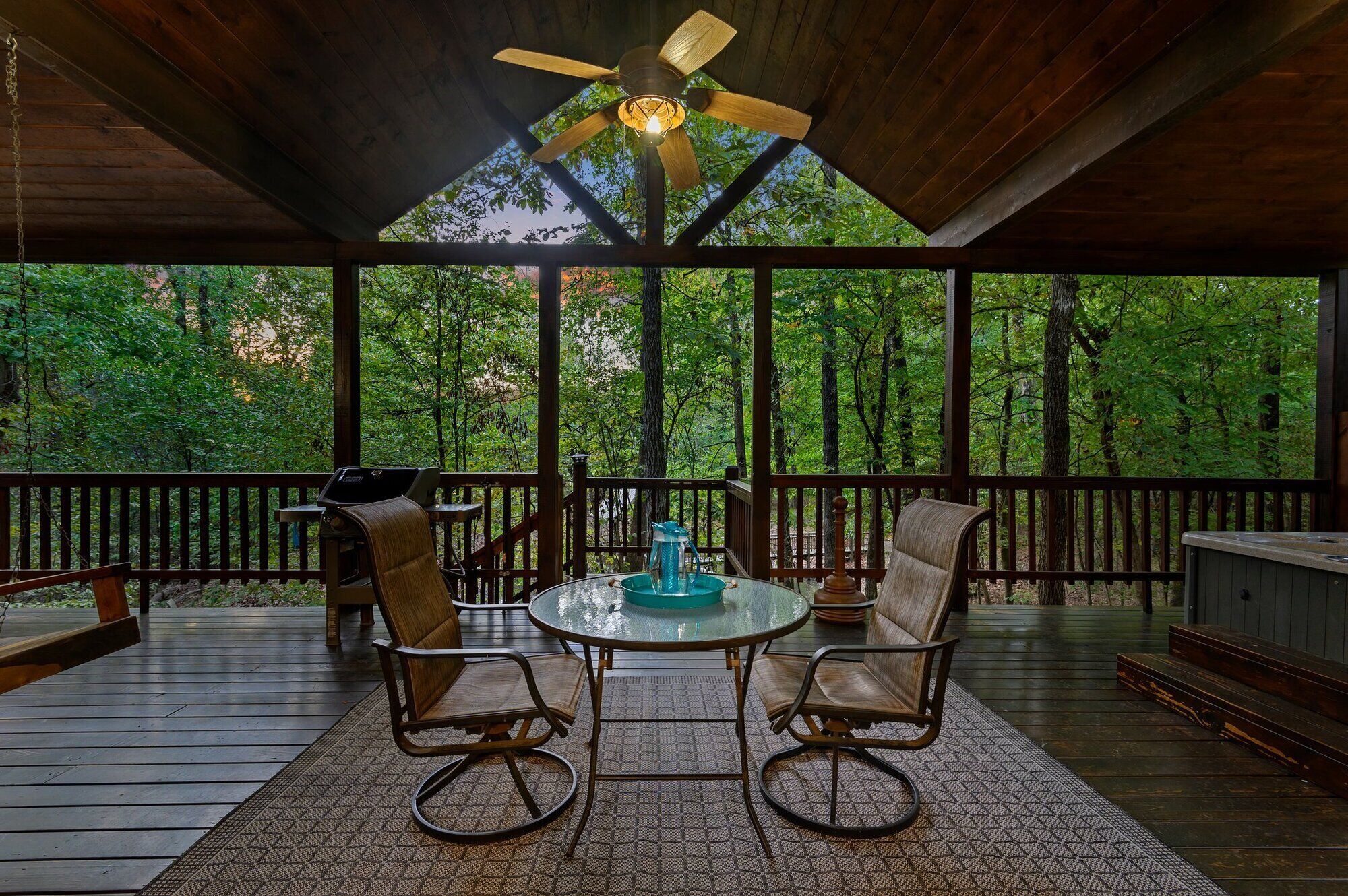 A covered outdoor seating area with a table and chairs, a ceiling fan, and a view of a forested area.