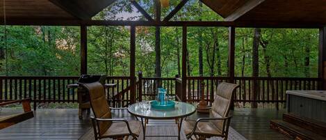 A covered outdoor seating area with a table and chairs, a ceiling fan, and a view of a forested area.