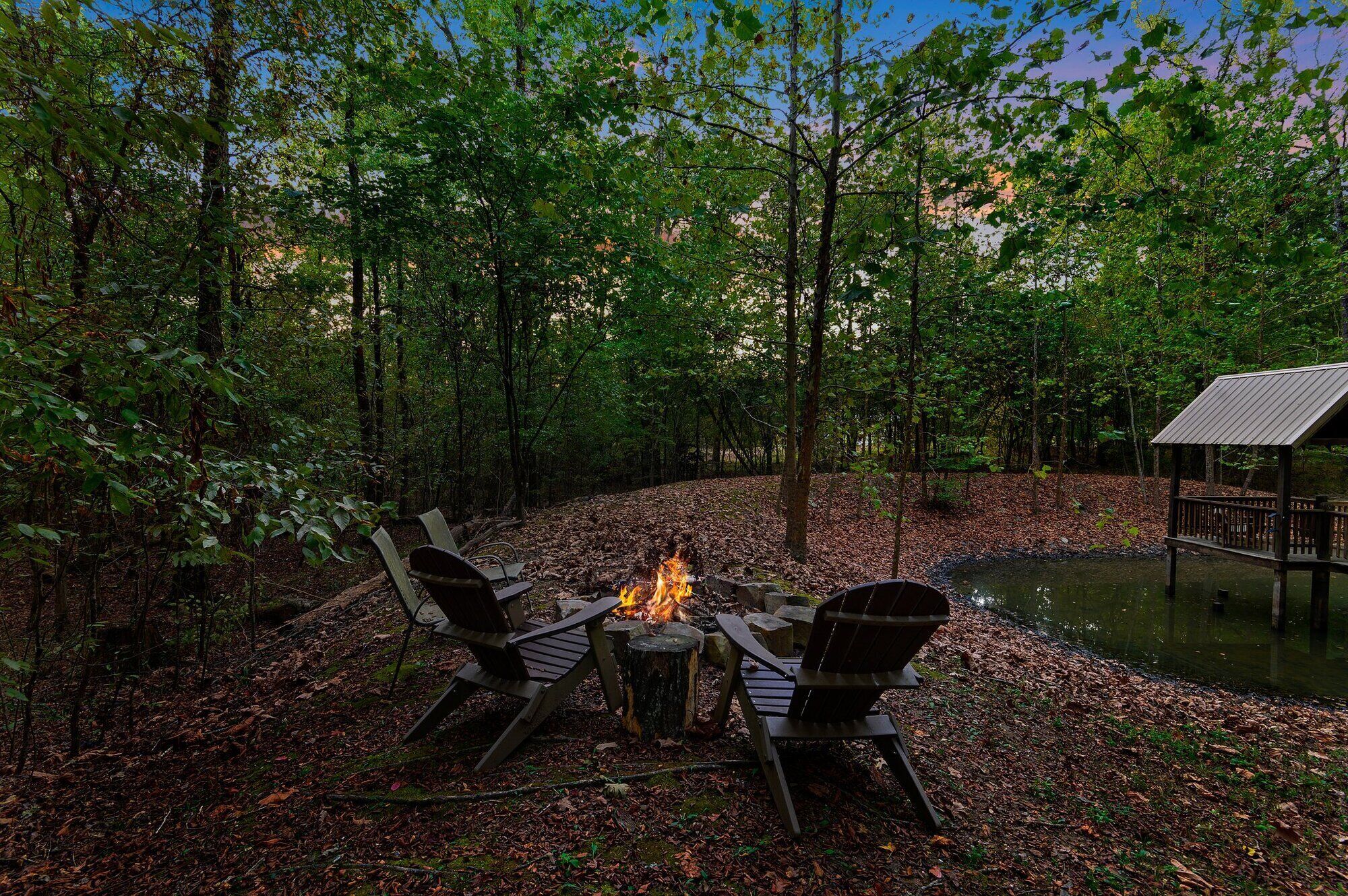 Un feu de camp, avec deux chaises Adirondack placées en face, entouré d’une zone boisée comprenant une terrasse en bois et un étang.