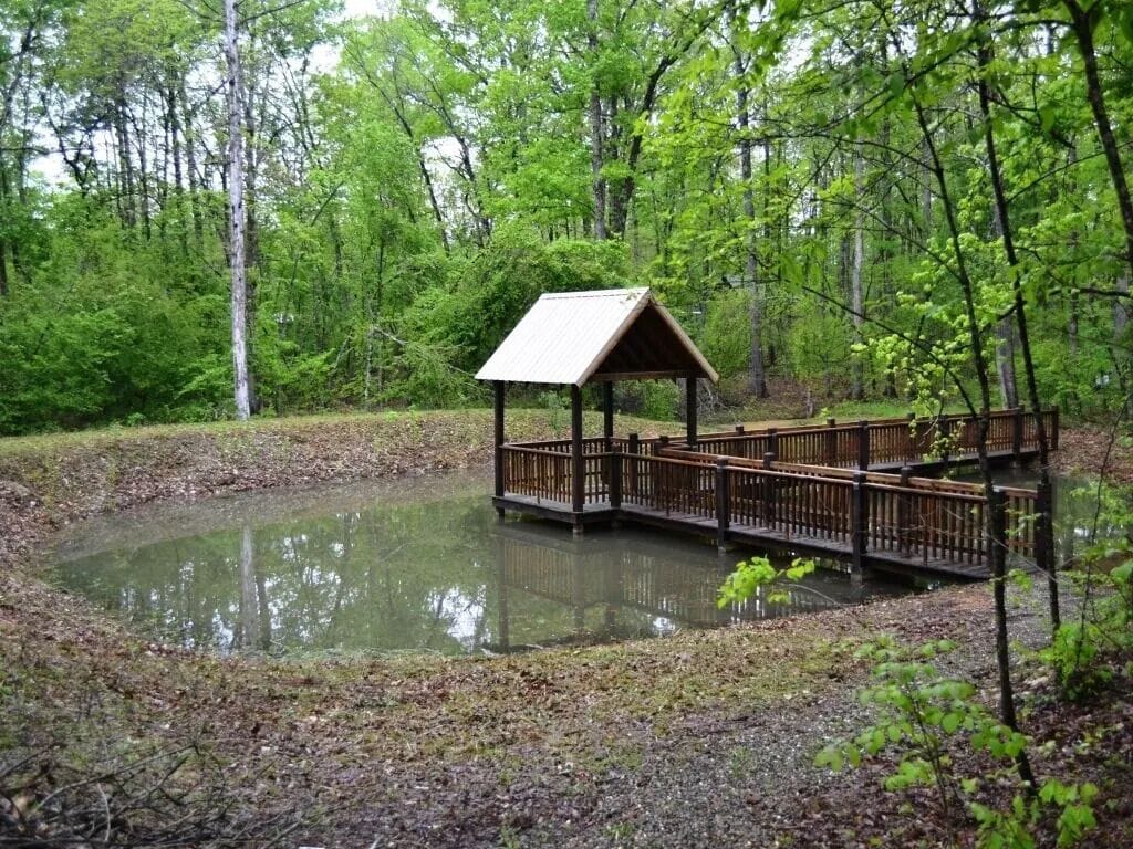 Ein hölzerner Pavillon mit Metalldach über einem Teich, umgeben von Bäumen.