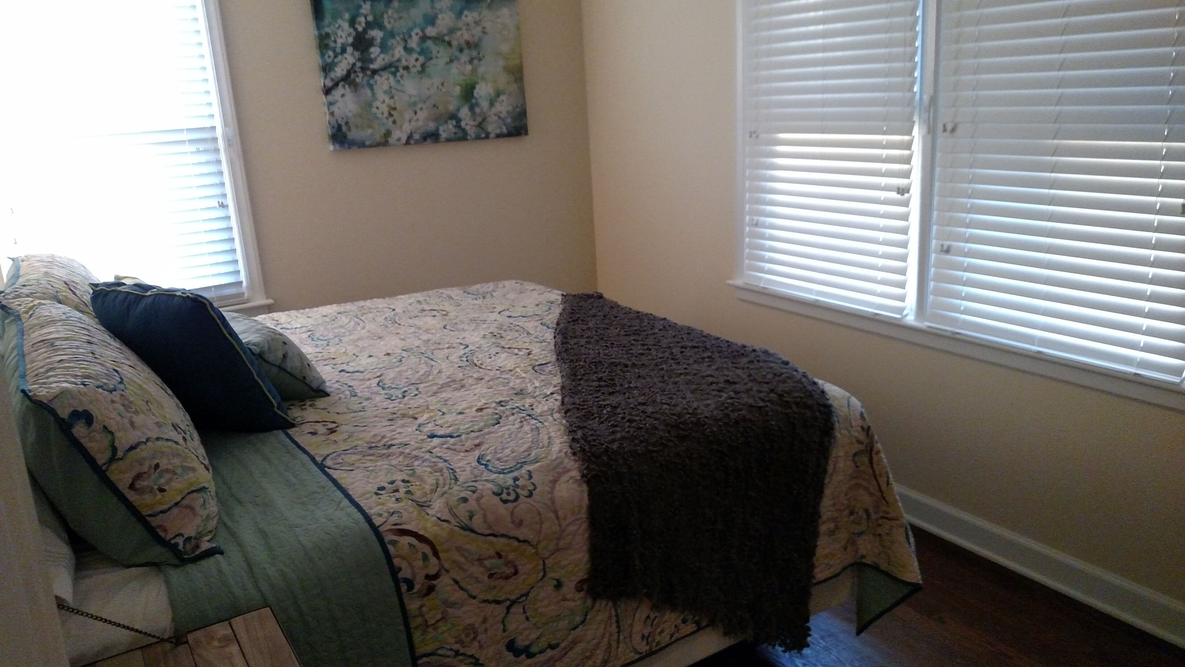A bedroom with a bed, a window with blinds, and a wall with a floral painting.