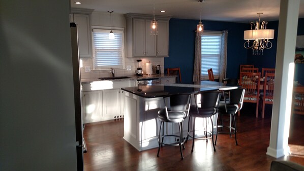 A kitchen with white cabinets, a central island with bar stools, a chandelier, and a dining area with wooden chairs. - Open Floor Plan 2 Bdrm-Private Home on Half Acre-Garage Parking (Mission)
