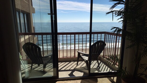 Ein Balkon mit zwei Stühlen und einem Tisch, der auf einen Strand mit dem Meer und einem wolkenlosen Himmel blickt.