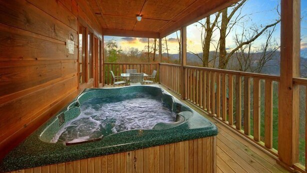 A wooden cabin with a hot tub on a deck, overlooking a mountainous landscape.