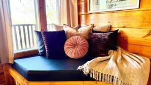A wooden bench with cushions and a blanket, a window with curtains, and a framed sign on the wall.