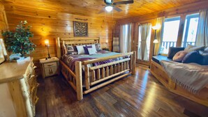 A wooden cabin bedroom with a bed, nightstands, a plant, and a window with curtains.