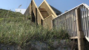 A wooden structure with a sloped roof, surrounded by tall grass and sand. - "Comes A Time" Oceanfront - Best Surf City location, walk to everything! (Surf City)