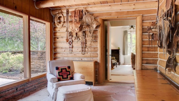 Interior de cabana de madeira com lareira de pedra, uma poltrona acolchoada e parede decorada com artigos de couro.