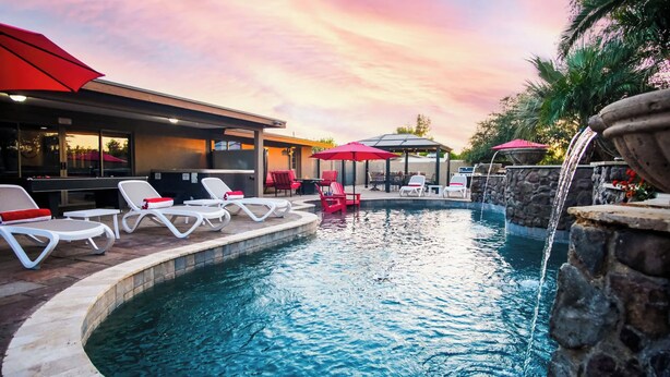 A pool area with lounge chairs and umbrellas, a stone wall with a water feature, and a building with large windows.