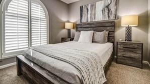 A bedroom with a large bed, two bedside tables, a headboard, and a window with blinds.