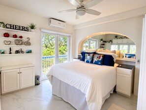A bedroom with a bed, a ceiling fan, a window with a view, and a shelf with decorative items.
