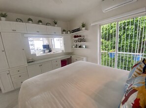 A bedroom with a large bed, a window with a view of greenery, and a kitchen area with cabinets and a TV.