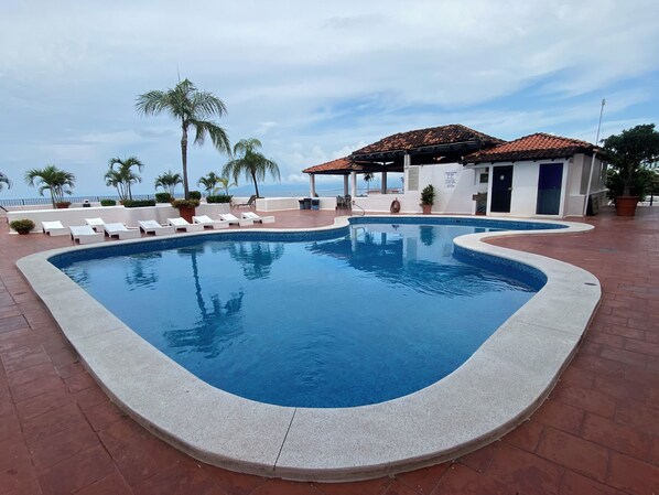 A swimming pool with lounge chairs and palm trees, a covered seating area, and a view of the ocean.