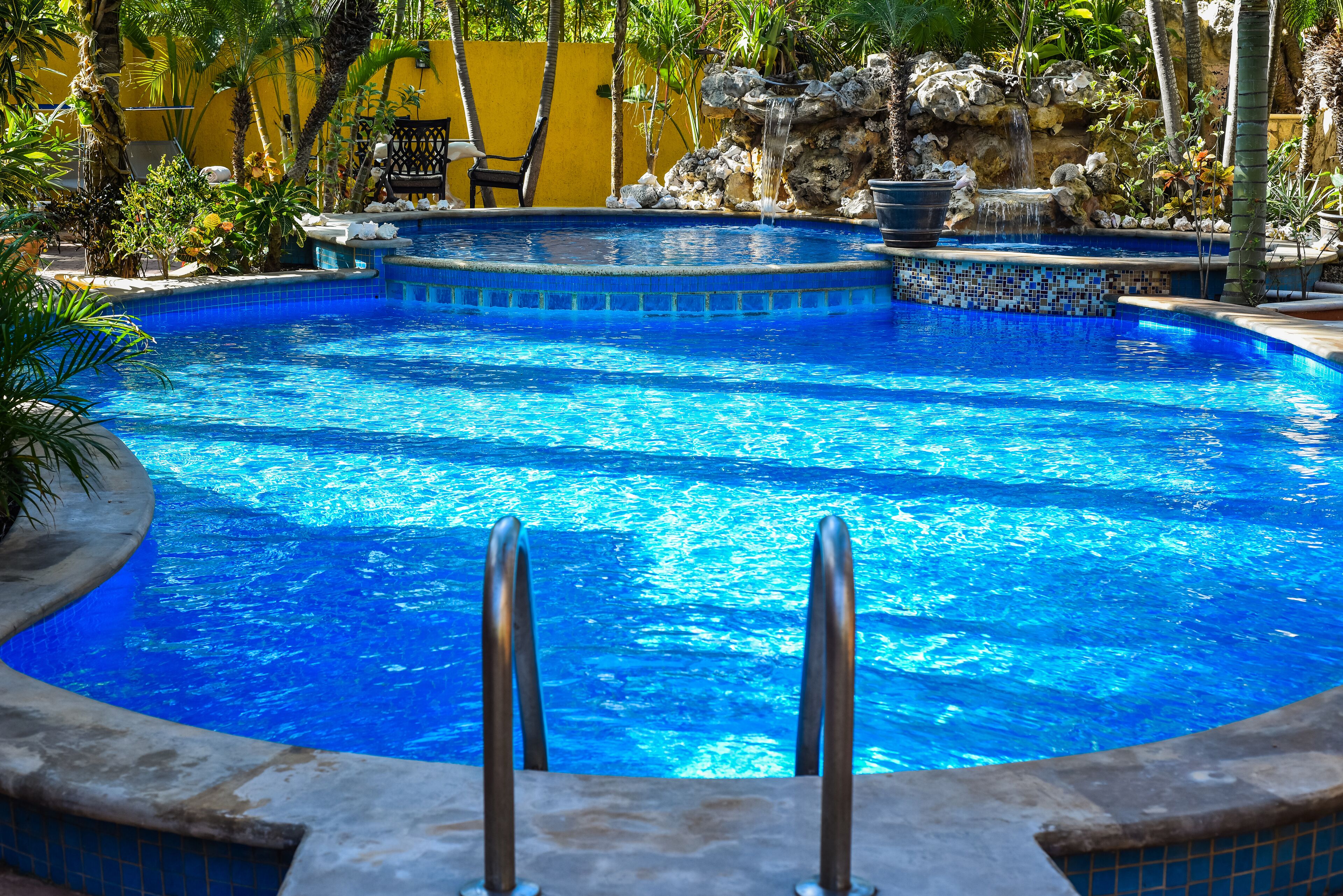 A clear blue swimming pool with a stone edge, a small waterfall feature, and a chair by the poolside.