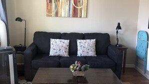 A living room with a dark sofa, patterned cushions, a coffee table with a floral centerpiece, a colorful abstract painting on the wall, and a blue chair. - Convenient & Cozy Cottage On Newport Near River, Downtown & Drake Park (Bend)