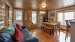 A log cabin interior with a wooden ceiling, a sofa with red cushions, a dining table with chairs, and a window with a view of trees.