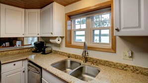 A kitchen with two stainless steel sinks, a granite countertop, wooden ceiling, and a window with a view of trees.