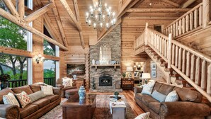 A log cabin interior with a stone fireplace, wooden staircase, and large windows. - Curated, One of a kind 7,000 sqft. Lodge, VIEWS & Pool (Sevierville)