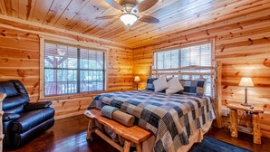 A log cabin bedroom with a bed, a leather chair, a bench, and a ceiling fan. - Relax by Broken Bow Lake, your cozy 3-bedroom Hochatown retreat (Broken Bow)