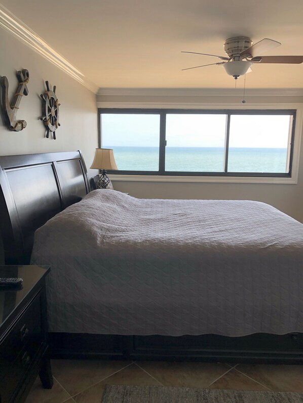 A bedroom with a large bed, a ceiling fan, a window with a view of the sea, and wall decor.