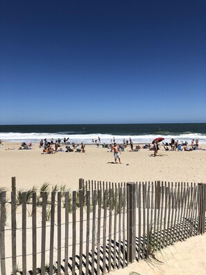 A beach with people sunbathing and swimming, a wooden fence in the foreground, and the ocean in the background.