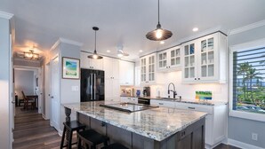 A modern kitchen with white cabinets, a large island with stools, pendant lighting, and a view through the window. - The Dream View Vacation - OCEAN FRONT -  (Princeville)