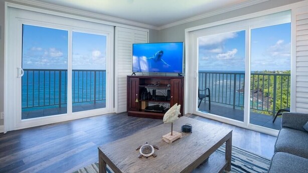 A living room with a large TV, wooden coffee table, and a view of the ocean through sliding glass doors. - The Dream View Vacation - OCEAN FRONT -  (Princeville)
