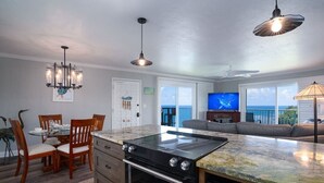 A modern kitchen with a large island, wooden chairs, a dining table, and a view of the ocean. - The Dream View Vacation - OCEAN FRONT -  (Princeville)