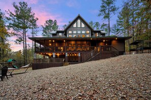 A large, two-story log cabin with a wrap-around porch and multiple windows. - Hot tub, Playground, 2 Fireplaces- Great for Large Families (Broken Bow)