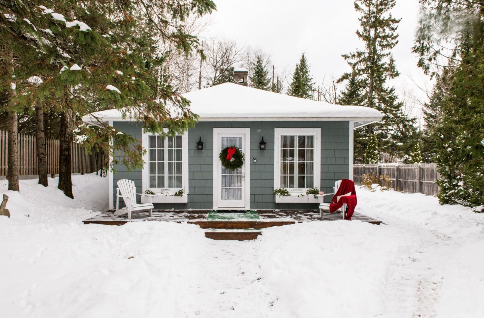 A house with a snow-covered roof, a front porch with a wreath, and a wooden fence in the background.