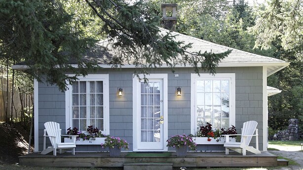 A small house with two white doors, a covered porch with two white Adirondack chairs, and potted plants.